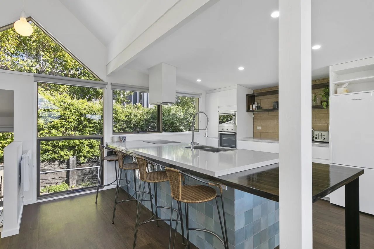 A bright kitchen with a long island and large windows overlooking trees.