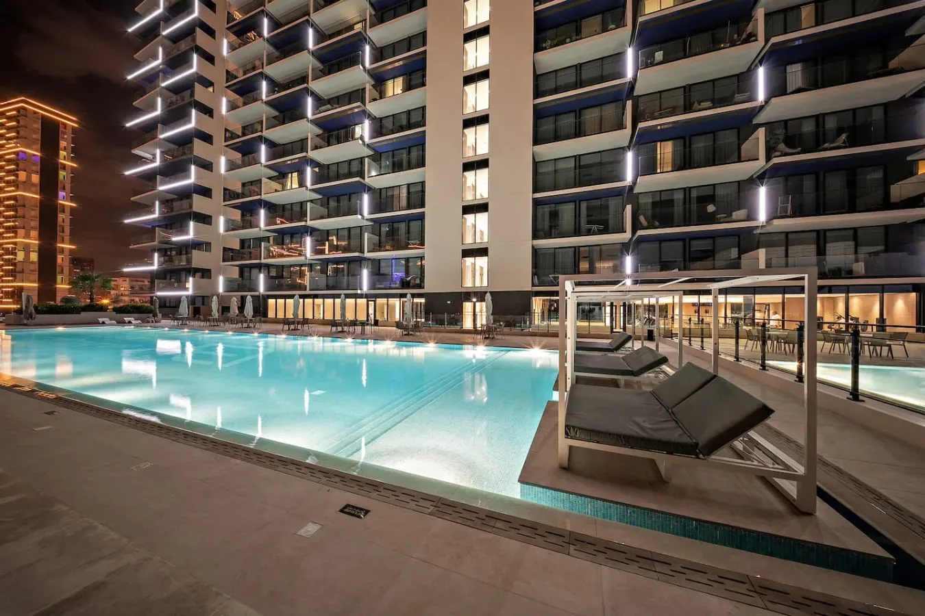 Una piscina exterior iluminada junto a un edificio moderno de gran altura por la noche.