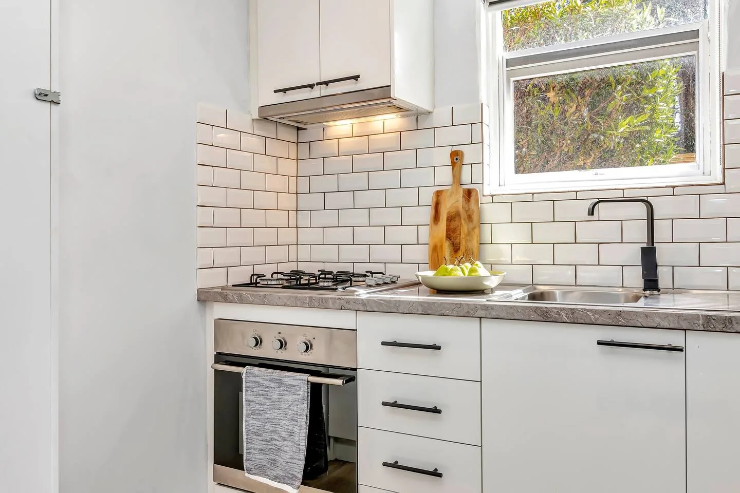 Una cocina luminosa tiene gabinetes blancos, un salpicadero de azulejos y una ventana sobre la encimera.