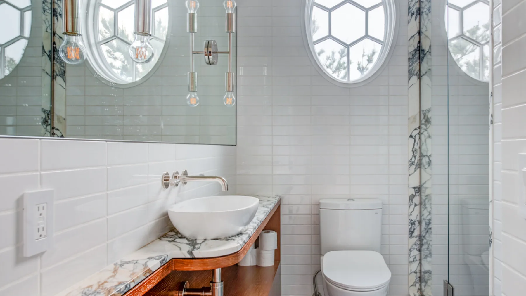Un baño con azulejos tiene un lavabo sobre encimera y un inodoro bajo ventanas redondas.