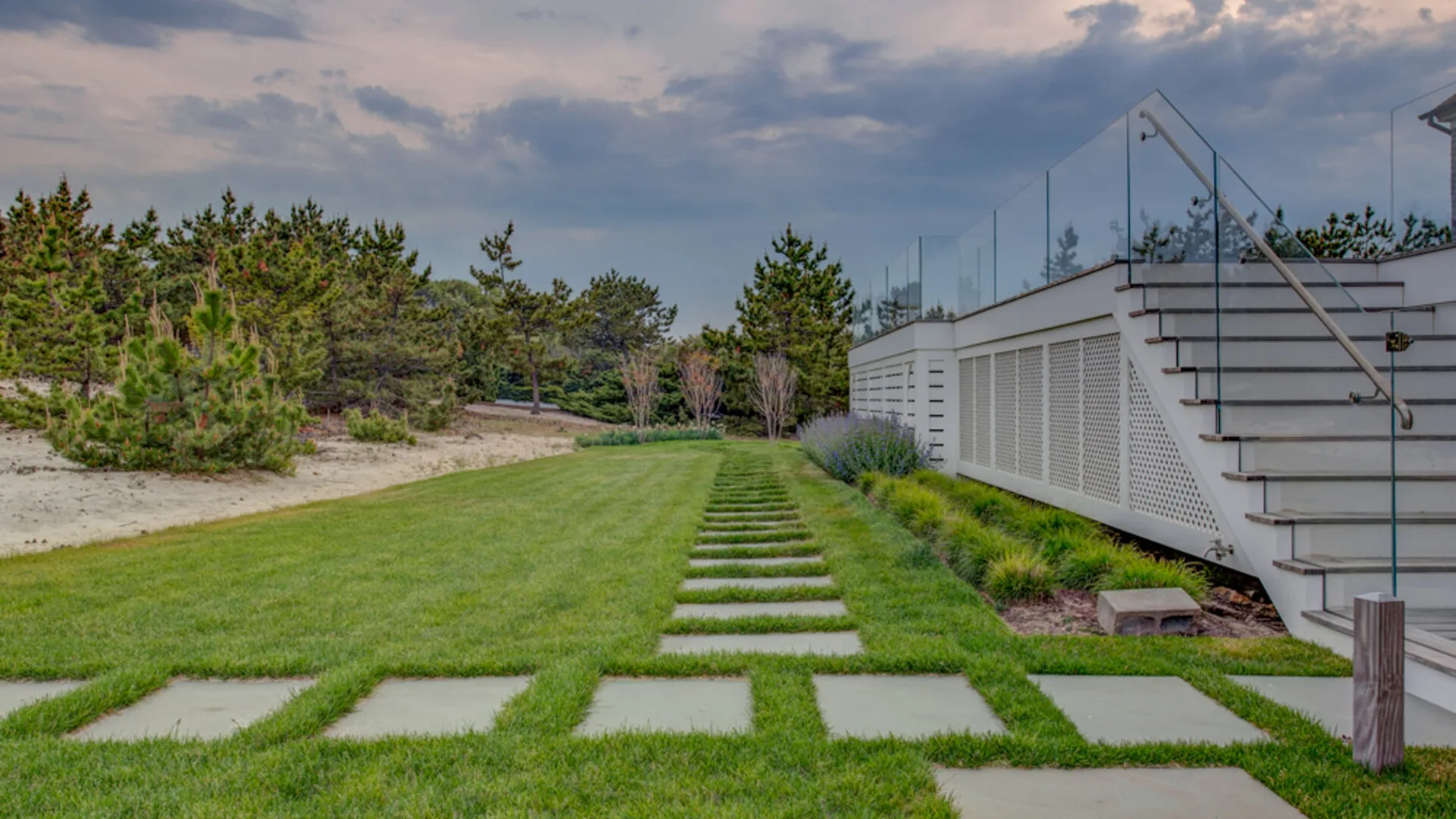Un sendero ajardinado conduce hacia un edificio moderno junto a una zona arenosa con árboles.