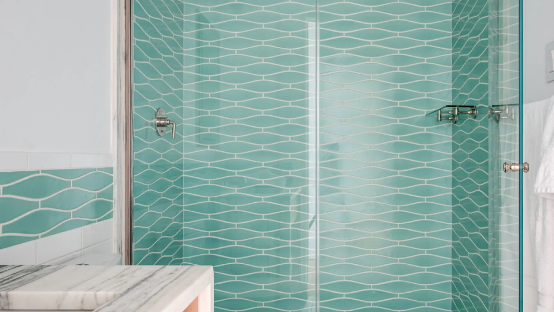 Una ducha de baño tiene paredes de azulejos turquesa con motivos y paneles de vidrio.