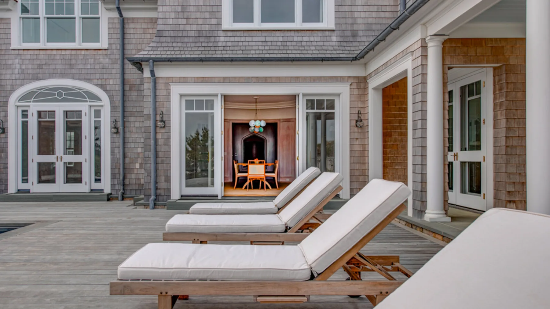 Una terraza de madera con asientos acolchados junto a una casa con revestimiento de tejas.