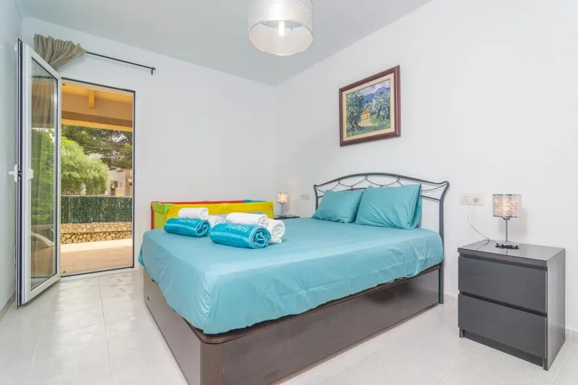 A bright bedroom with a bed dressed in turquoise linens and an open balcony door.