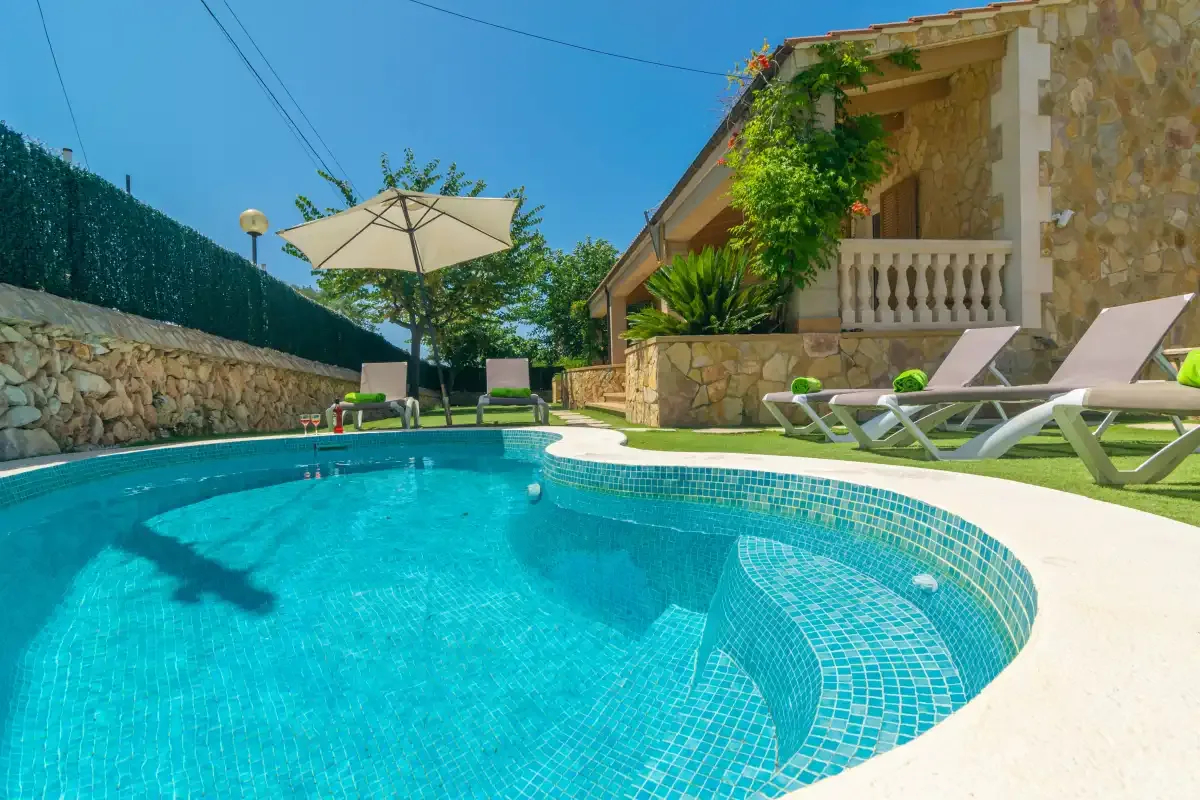 A swimming pool beside a stone house, with loungers and a patio umbrella.
