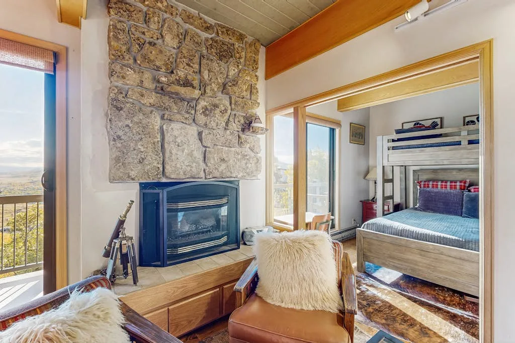 A bedroom with a stone fireplace and a bunk bed beside large windows.