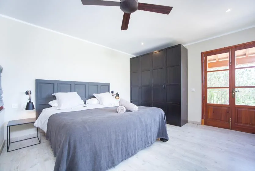 A bedroom with a neatly made bed, dark wardrobe, and wooden doors opening to a bright view.