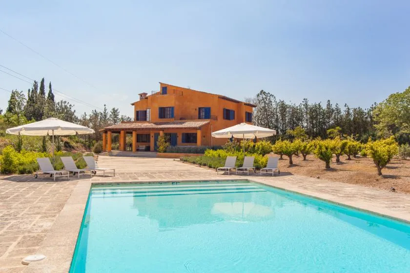 An outdoor swimming pool in front of an orange house, with loungers and umbrellas nearby.