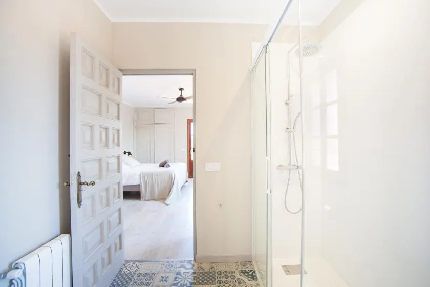 A bright bathroom with a glass shower beside an open doorway to a bedroom.