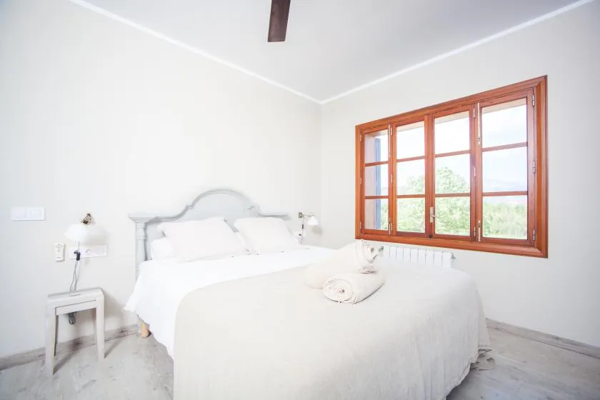 A bright bedroom with a white bed beside a large wood-framed window.