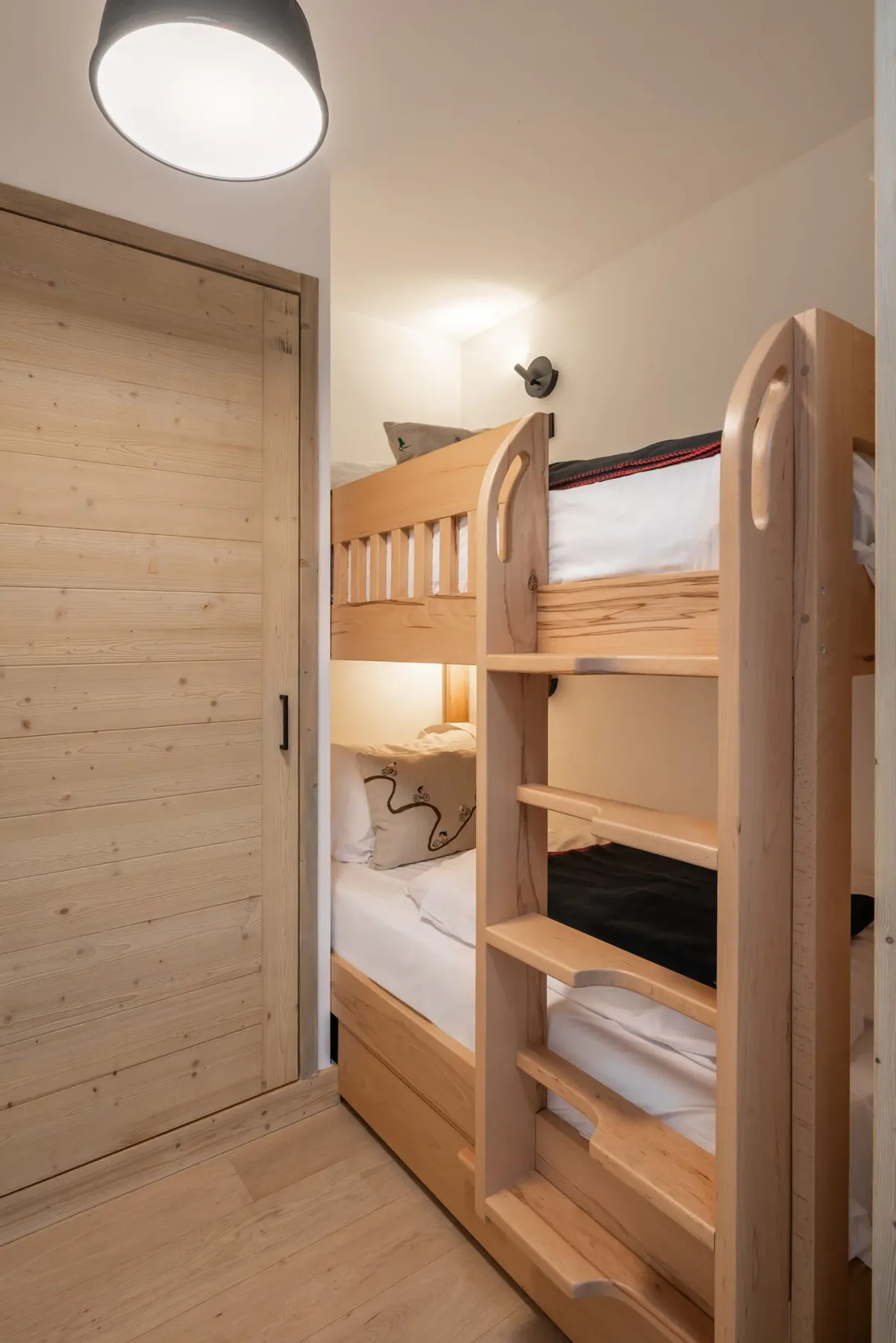 Narrow room with wooden bunk beds, a sliding wooden door, wall sconce lighting, and framed art on the wall.