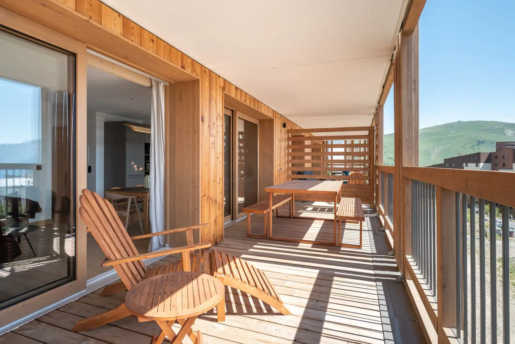 A wooden terrace with a lounge chair, a swing chair, and a small round table, overlooking a grassy landscape. A living room is visible through glass doors.