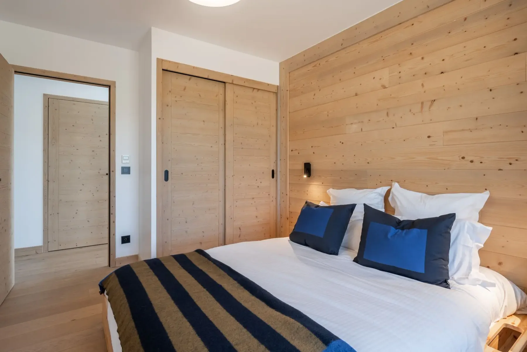 Cozy bedroom with wooden walls, a double bed adorned with blue and black striped bedding, and a window overlooking a scenic landscape.