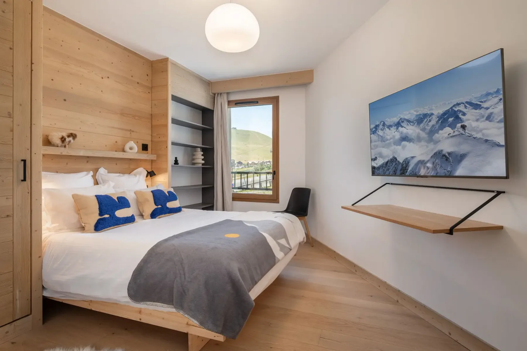 A cozy bedroom with a wooden accent wall, bed with white and blue pillows, wall-mounted TV, and a window view of mountains and greenery.