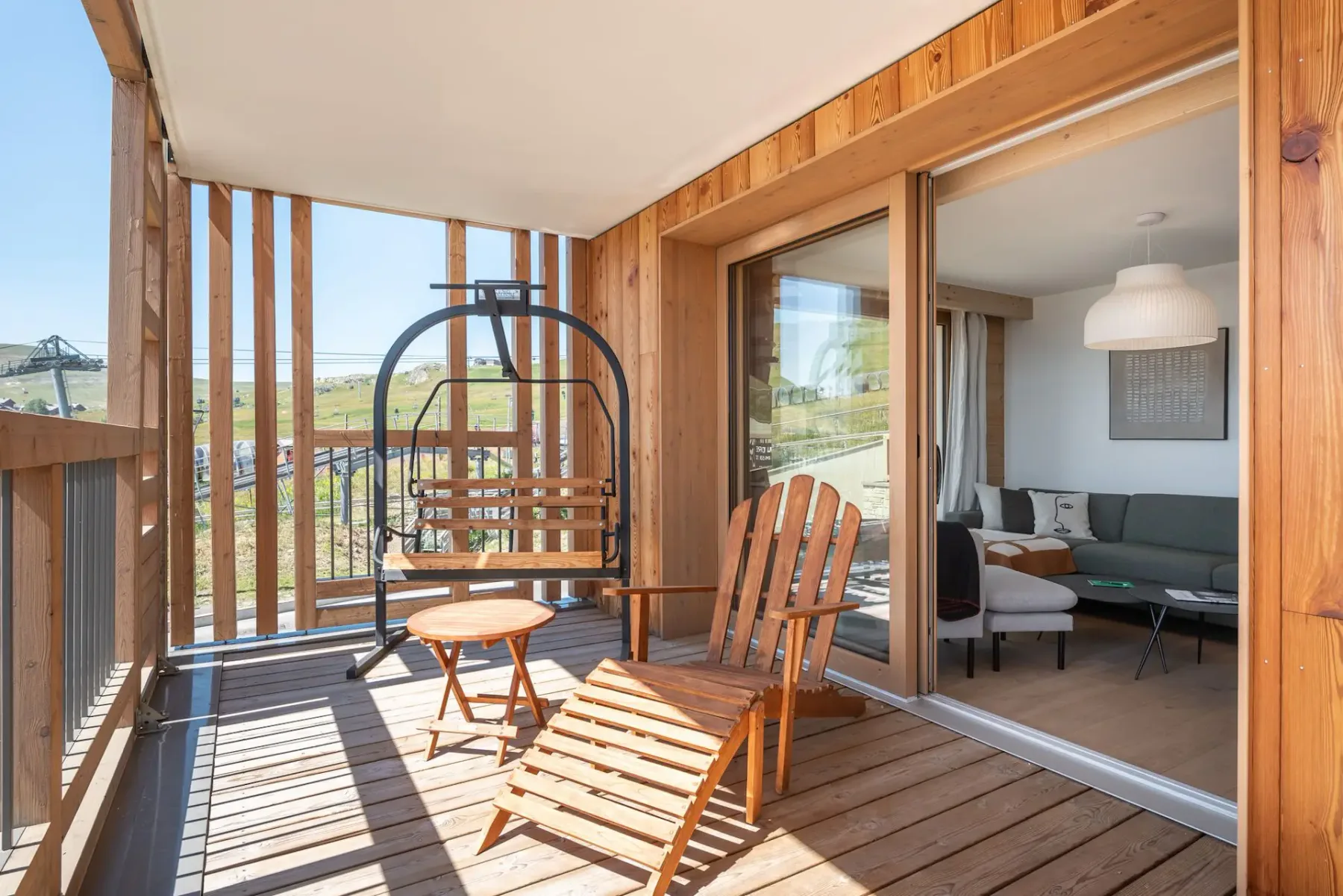 A wooden terrace with a lounge chair, a swing chair, and a small round table, overlooking a grassy landscape. A living room is visible through glass doors.