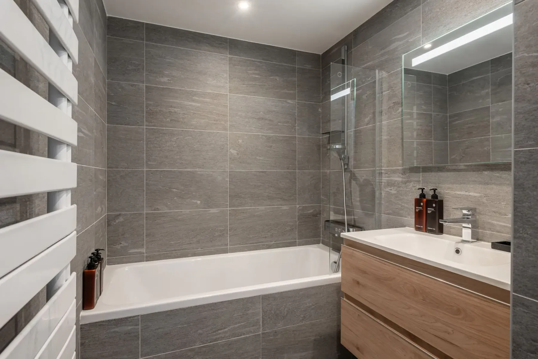 Modern bathroom with gray tiled walls, a bathtub, glass shower screen, wooden vanity with sink, and large mirror.