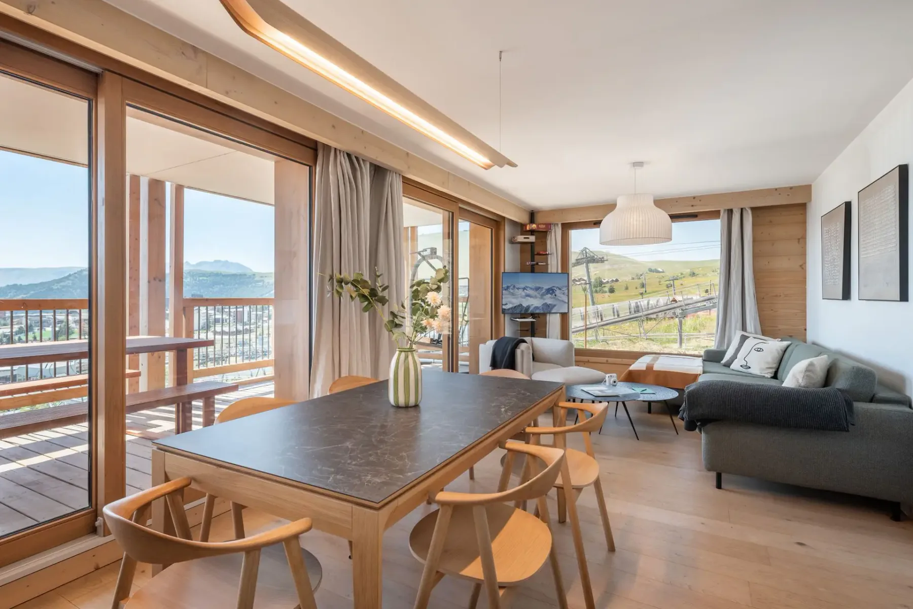 Modern kitchen with gray cabinets, wooden dining table, light wooden chairs, and a vase with greenery. Natural light from large windows brightens the room.