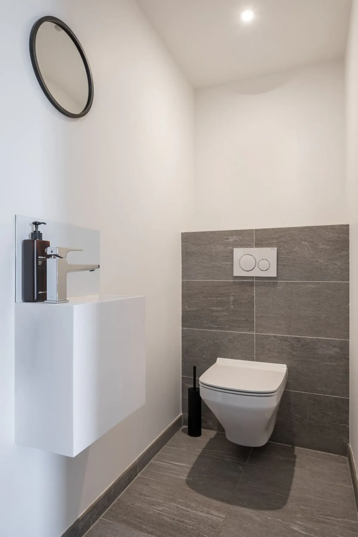 Minimalist bathroom with a wall-mounted toilet, gray tiles, a small sink with a soap dispenser, a round mirror, and overhead lighting.