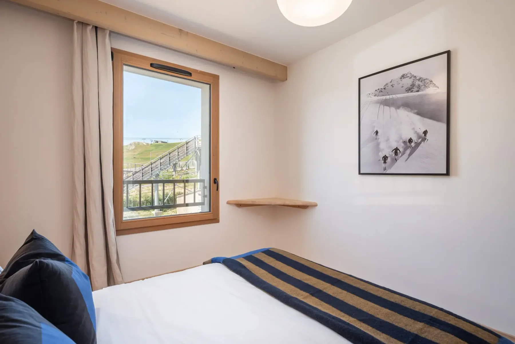 Cozy bedroom with wooden walls, a double bed adorned with blue and black striped bedding, and a window overlooking a scenic landscape.