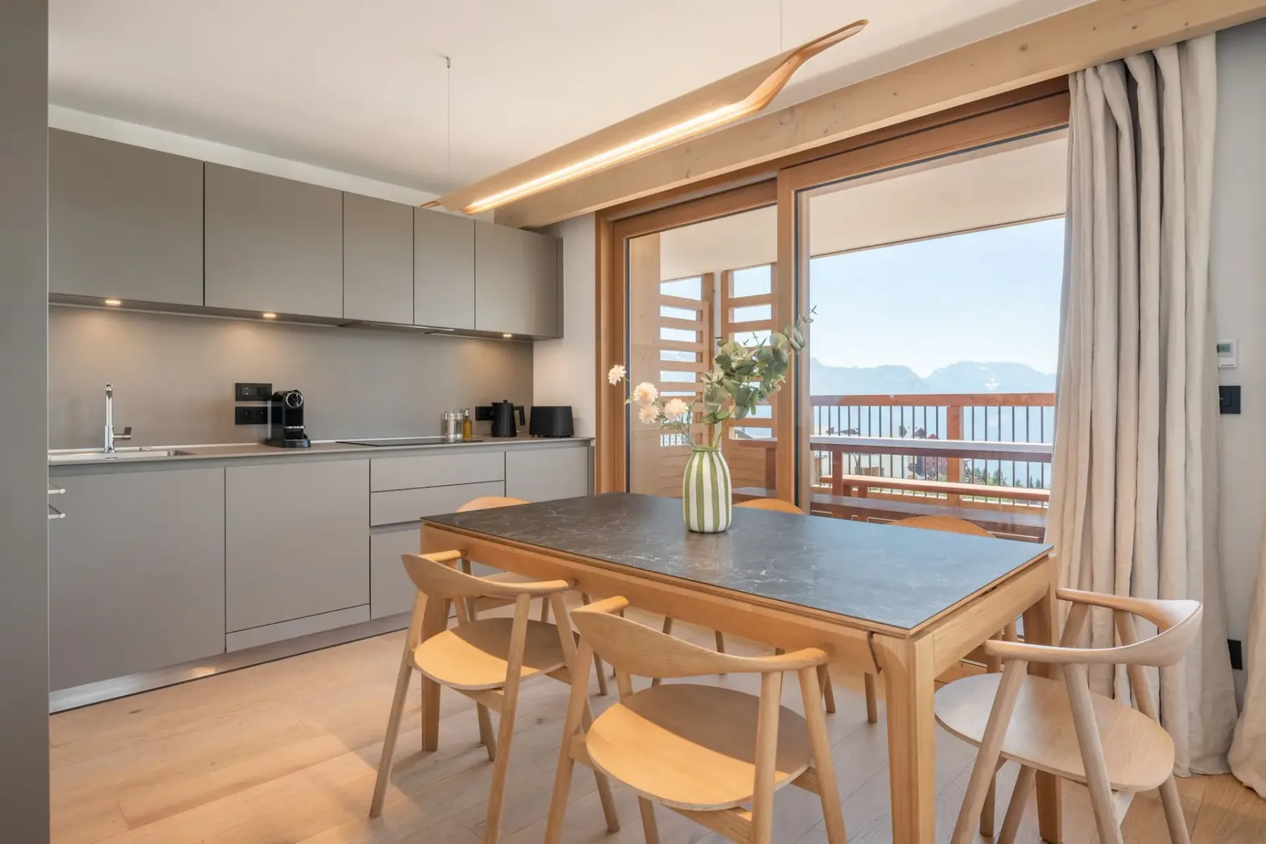 Modern kitchen with gray cabinets, wooden dining table, light wooden chairs, and a vase with greenery. Natural light from large windows brightens the room.
