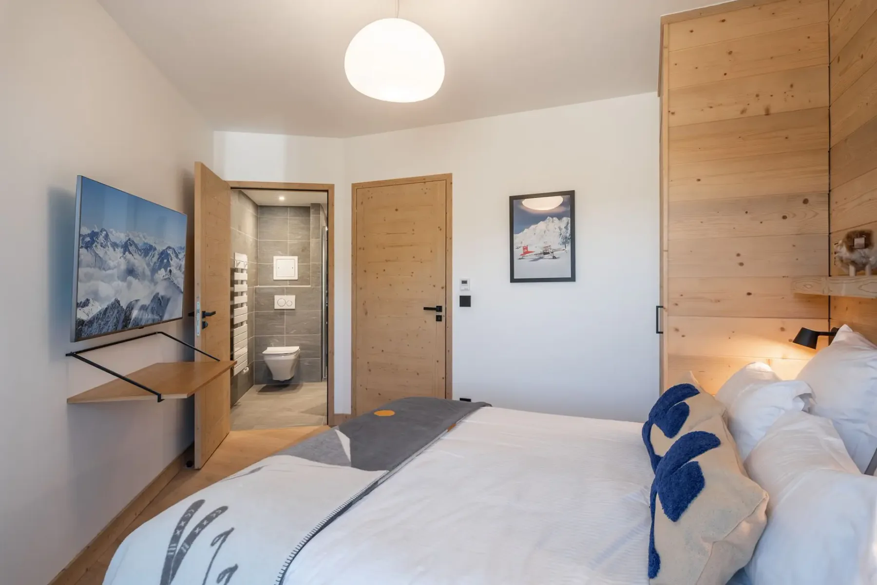 Cozy bedroom with a wooden accent wall, bed with decorative pillows, wall-mounted TV, and a view of mountains through the window.