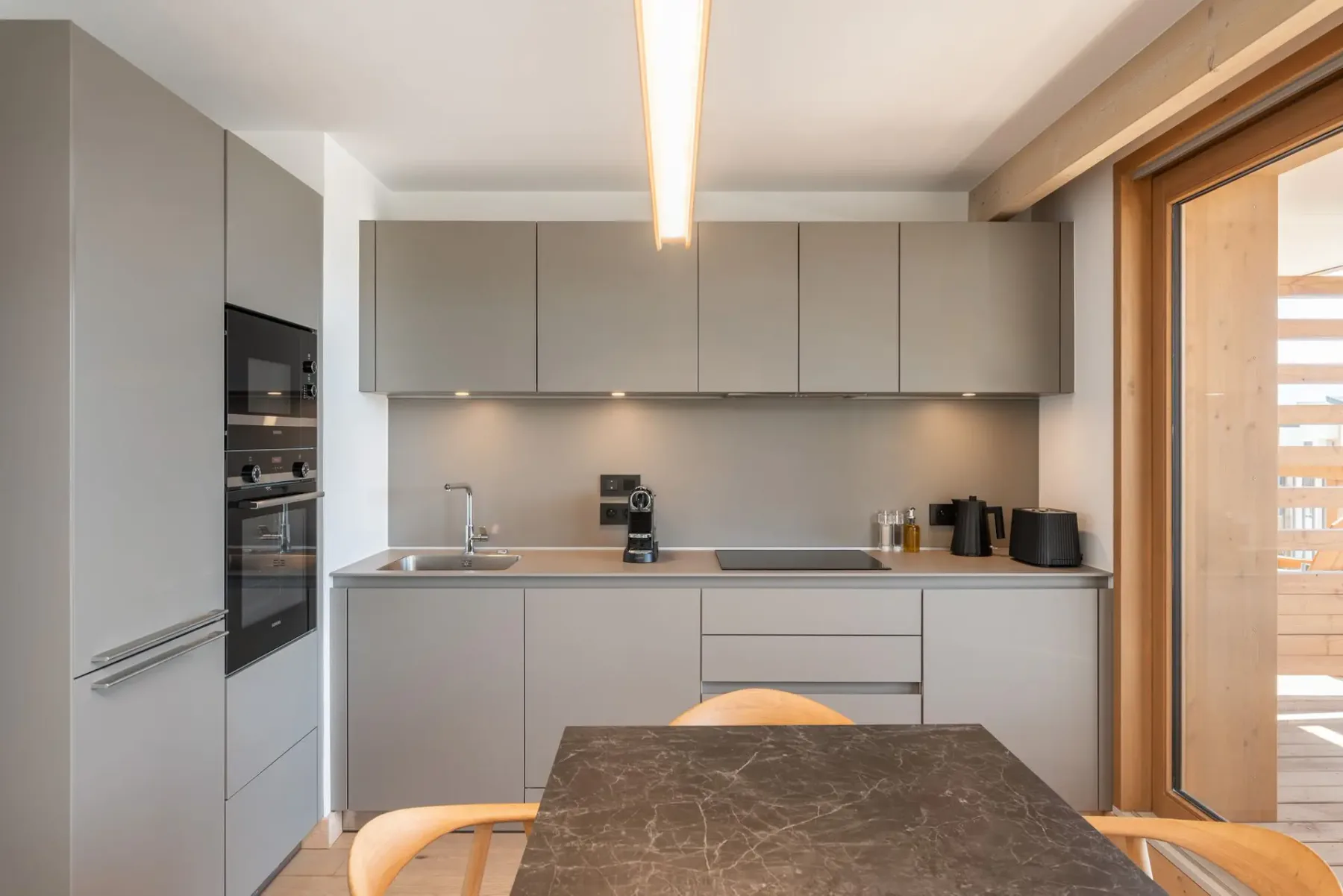Modern kitchen with gray cabinets, wooden dining table, light wooden chairs, and a vase with greenery. Natural light from large windows brightens the room.