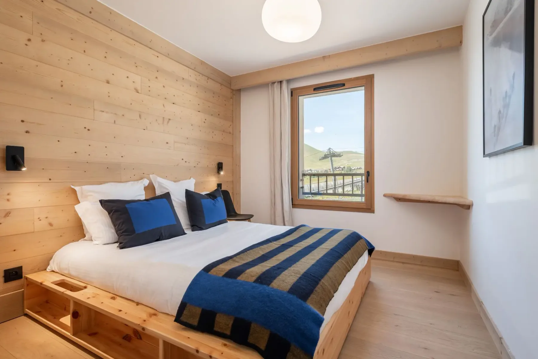 Cozy bedroom with wooden walls, a double bed adorned with blue and black striped bedding, and a window overlooking a scenic landscape.