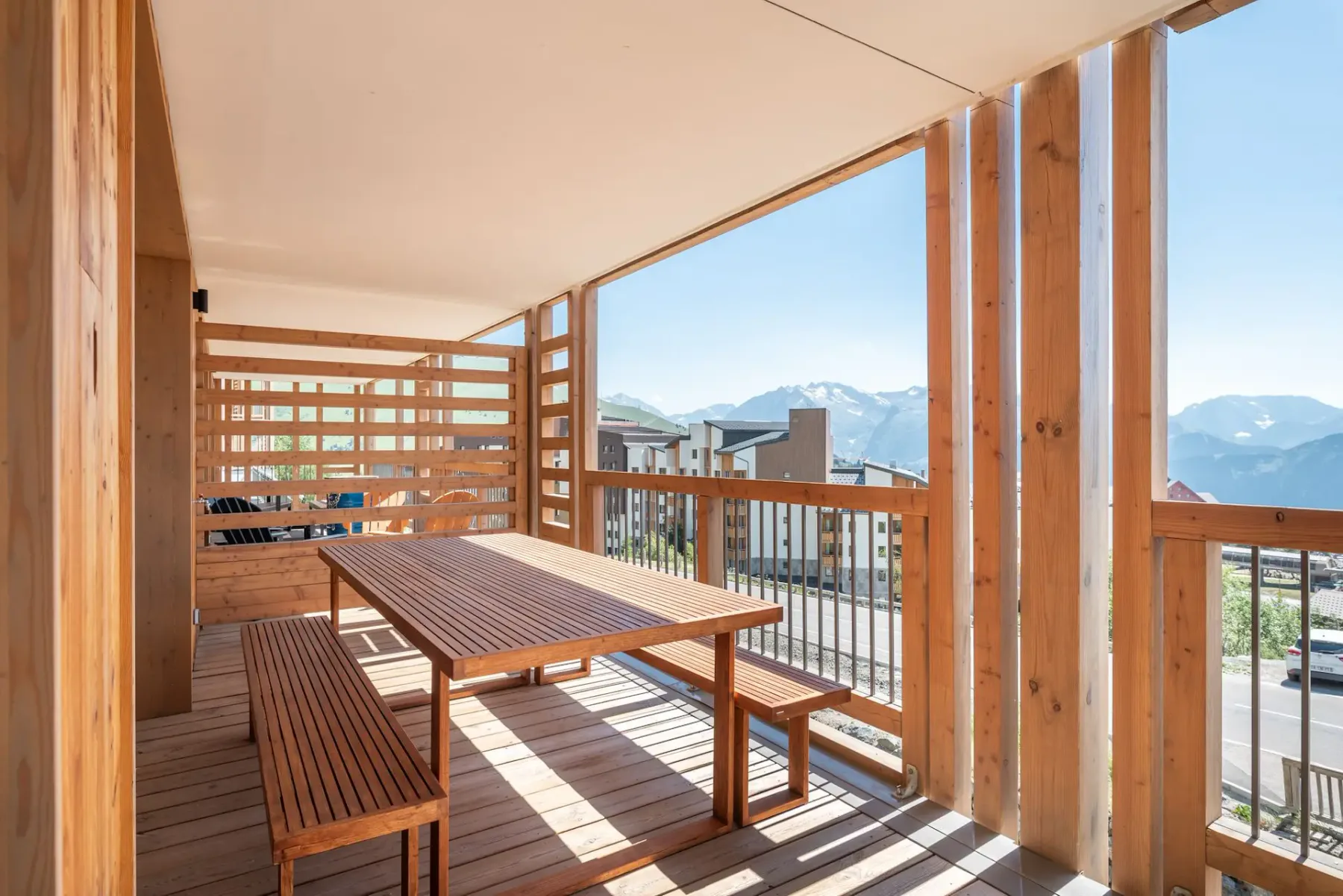 A wooden terrace with a lounge chair, a swing chair, and a small round table, overlooking a grassy landscape. A living room is visible through glass doors.