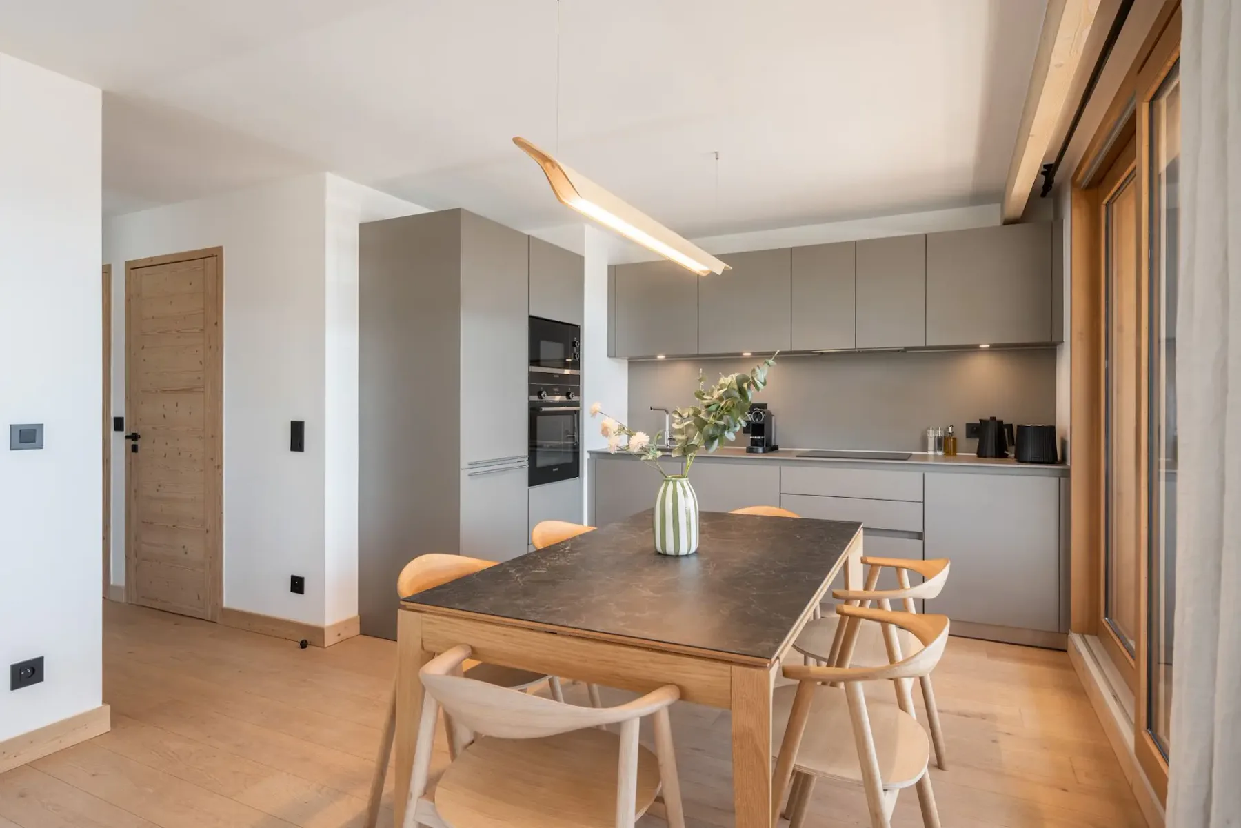 Modern kitchen with gray cabinets, wooden dining table, light wooden chairs, and a vase with greenery. Natural light from large windows brightens the room.