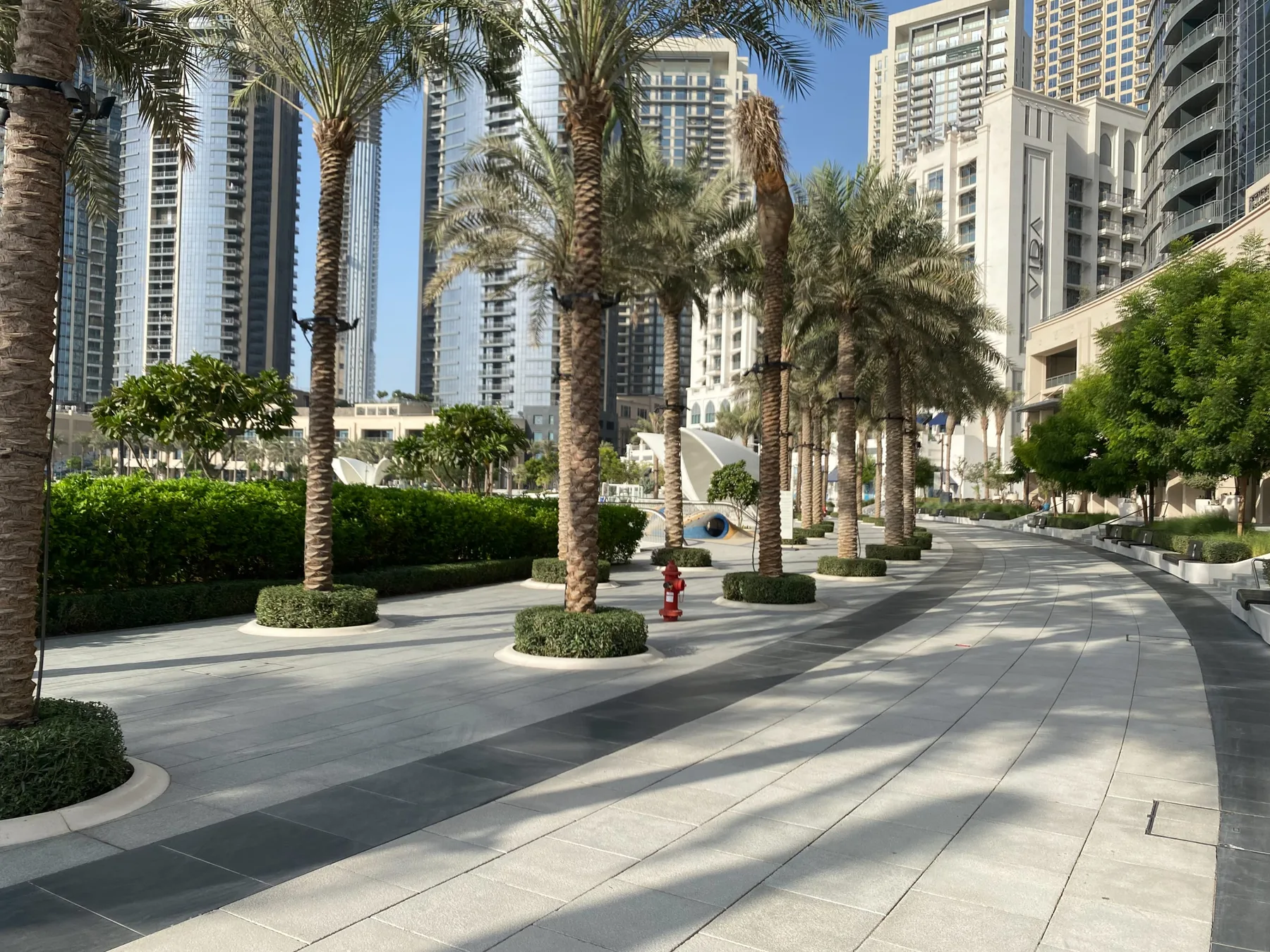 Un paseo peatonal pavimentado se curva entre palmeras y vegetación junto a edificios altos.
