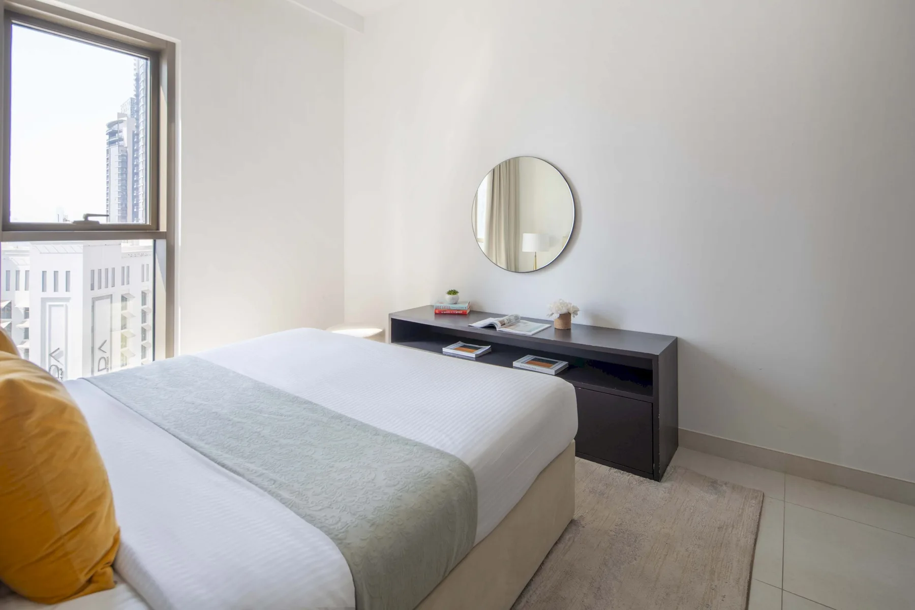 Un dormitorio luminoso con una cama tendida y un espejo redondo en la pared.
