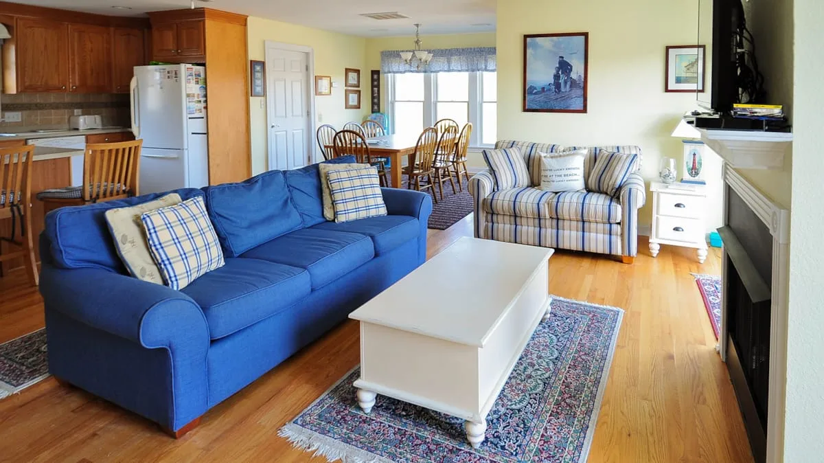 A living room with blue seating, a white coffee table, and a dining area beyond.