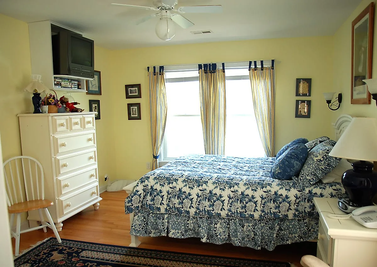 A bedroom with a bed covered in blue floral bedding beside bright curtained windows.