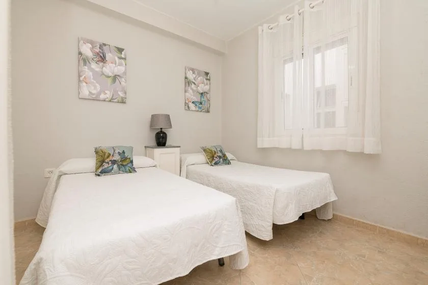 A bright bedroom with two beds and a window dressed with sheer curtains.