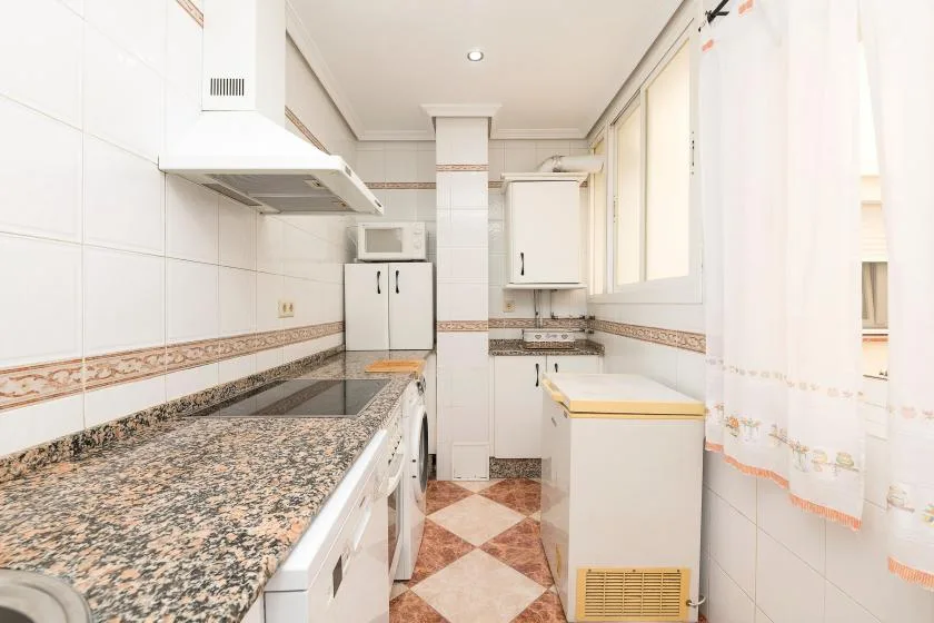 A narrow tiled kitchen with a long counter, cabinets, and a washing machine.