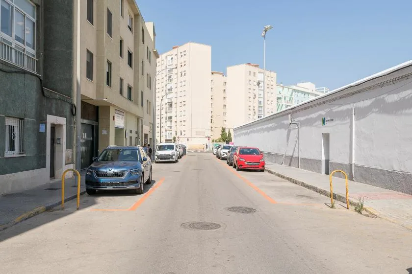 A sunny urban street runs between apartment buildings and parked cars.
