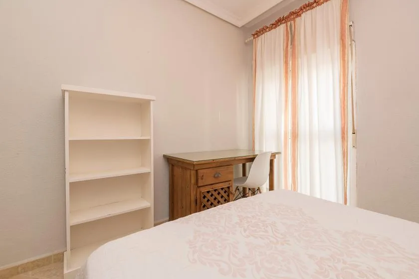 A bedroom with a bed, a desk, and a shelving unit beside a curtained window.