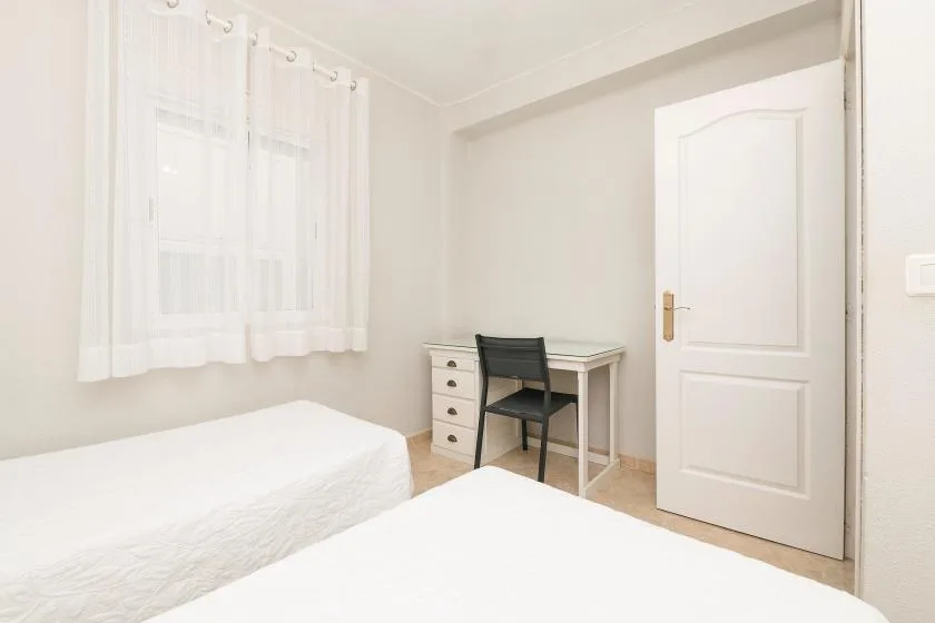 A bright bedroom with two beds and a small desk beside the window.