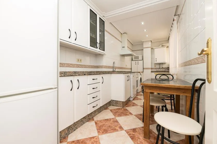 A narrow kitchen with white cabinetry, a tiled floor, and a wooden dining table.