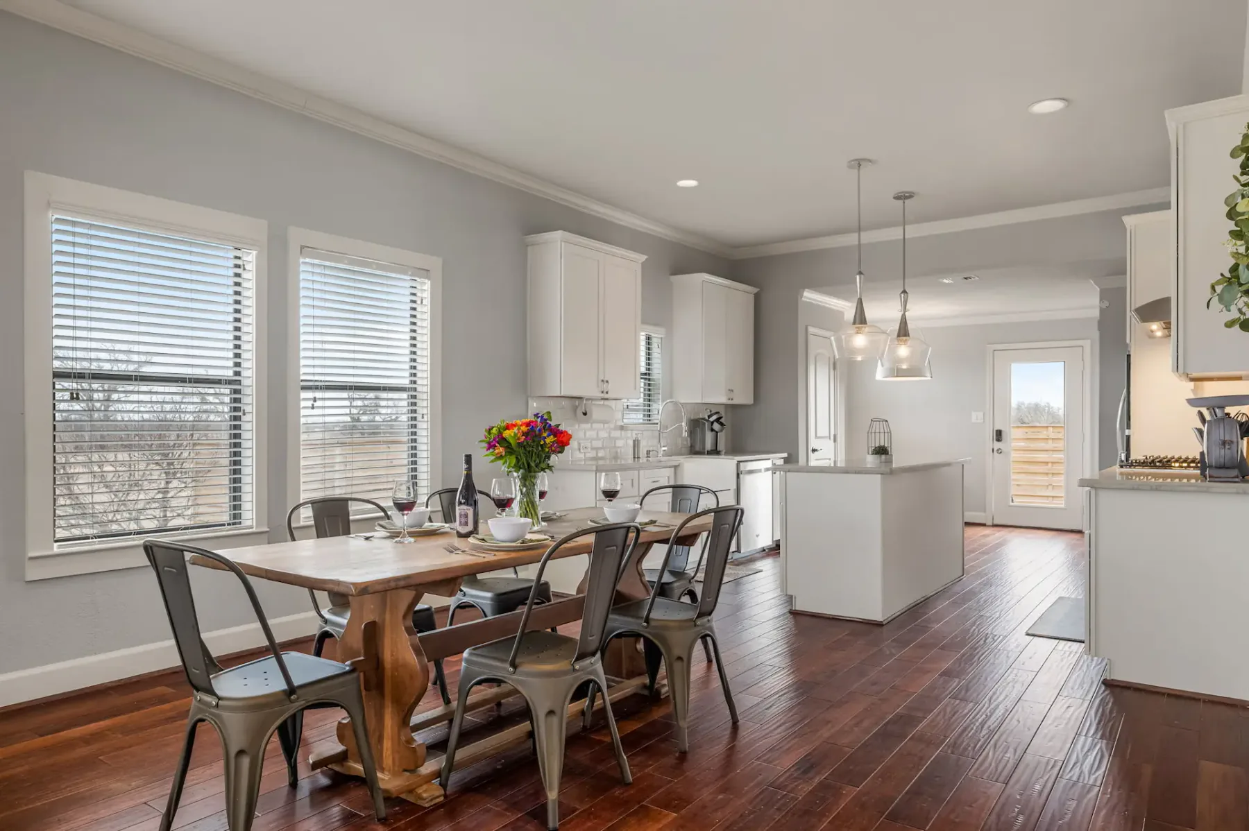 A modern kitchen with a wooden dining table, metal chairs, and pendant lights. Large windows allow natural light, emphasizing the hardwood floor.
