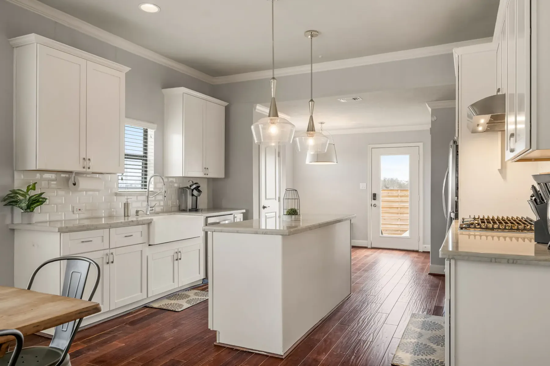 Modern kitchen with white cabinets, a central island, pendant lights, and wood flooring, leading to a sunlit door with an outdoor view.