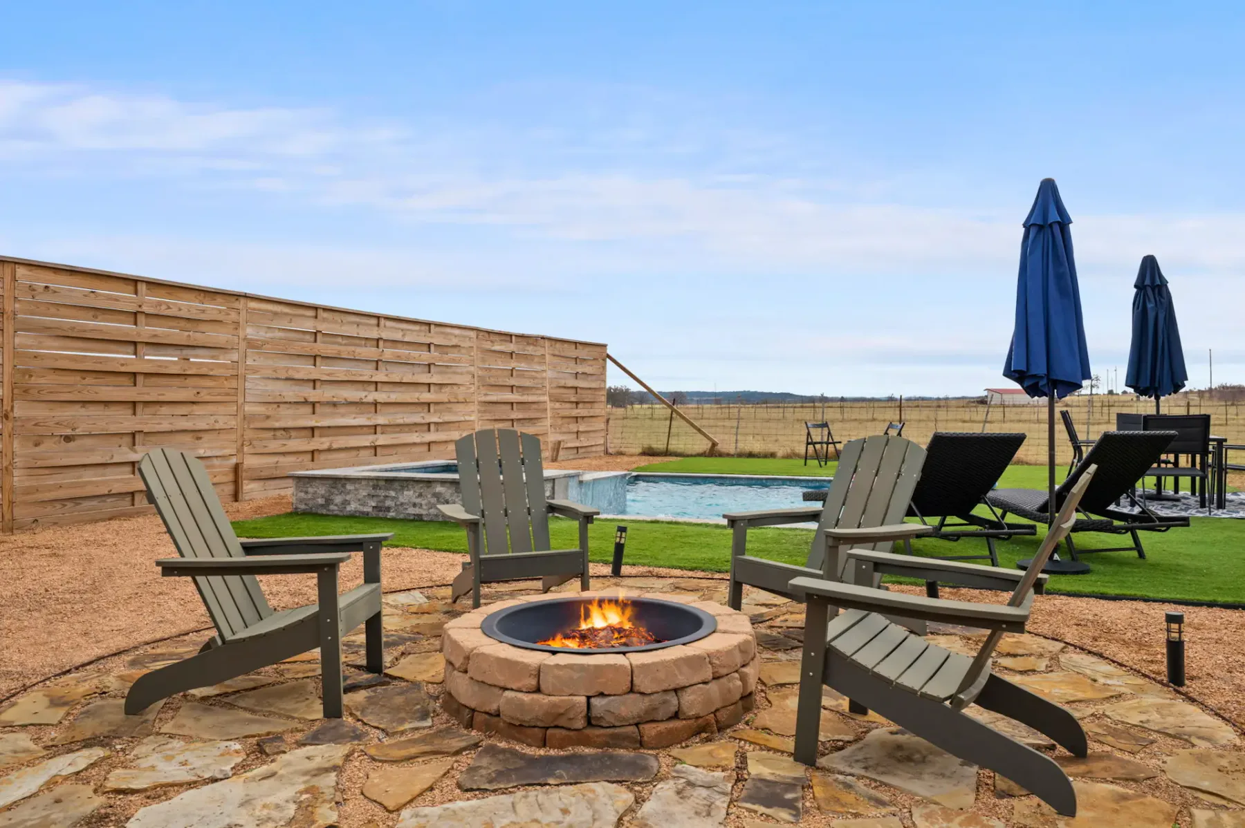 An outdoor patio with seating surrounds a lit stone fire pit beside a swimming pool.