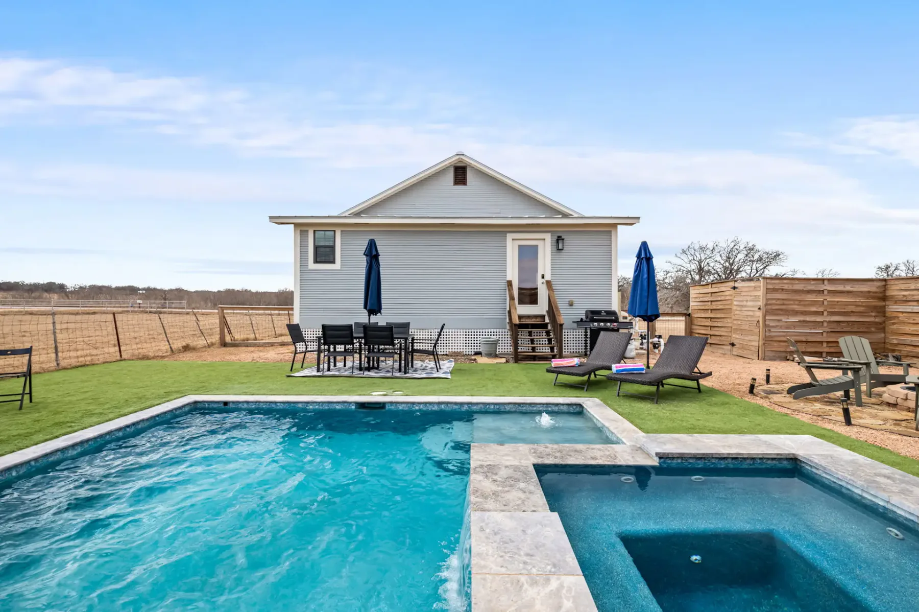 Backyard with a pool and hot tub, patio furniture, umbrellas, and a small house in the background, set in a rural landscape.