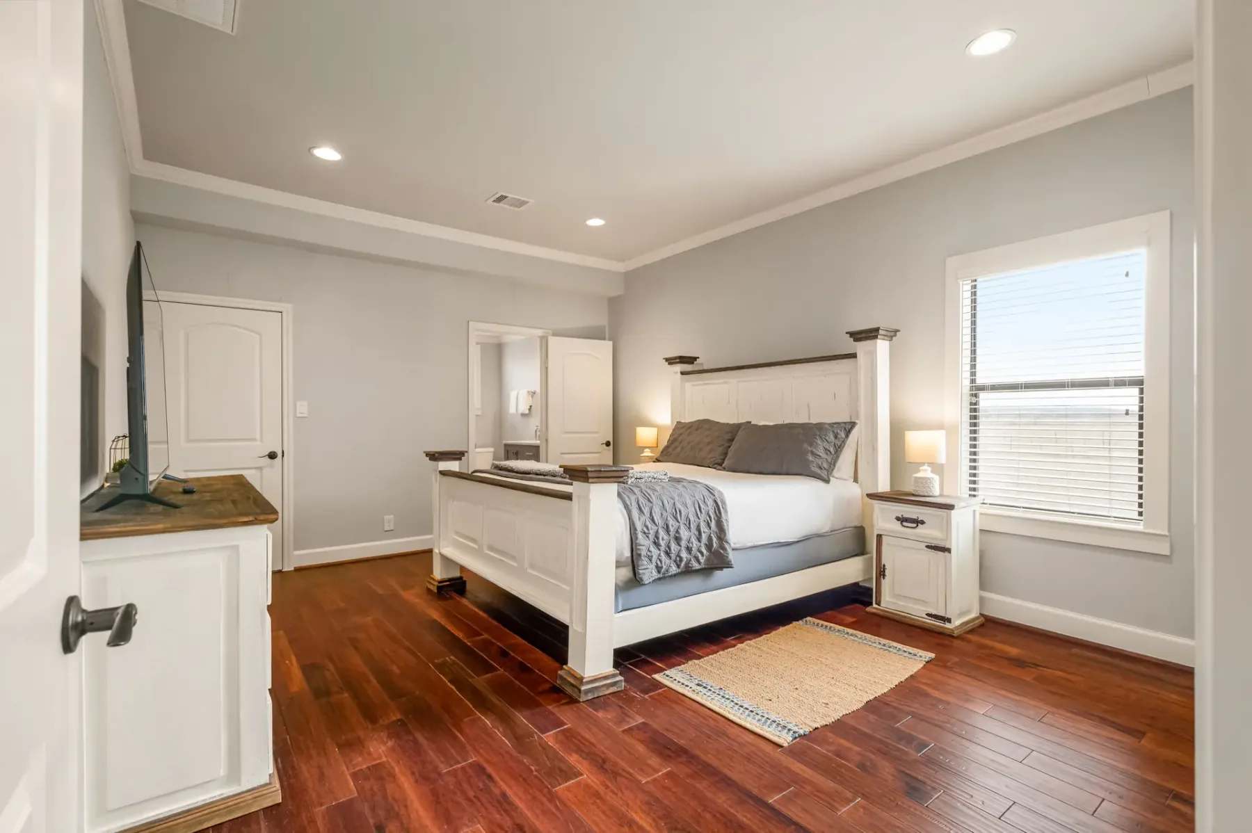 A modern bedroom with a large bed, hardwood floors, white and gray decor, a window, and a small rug on the floor.
