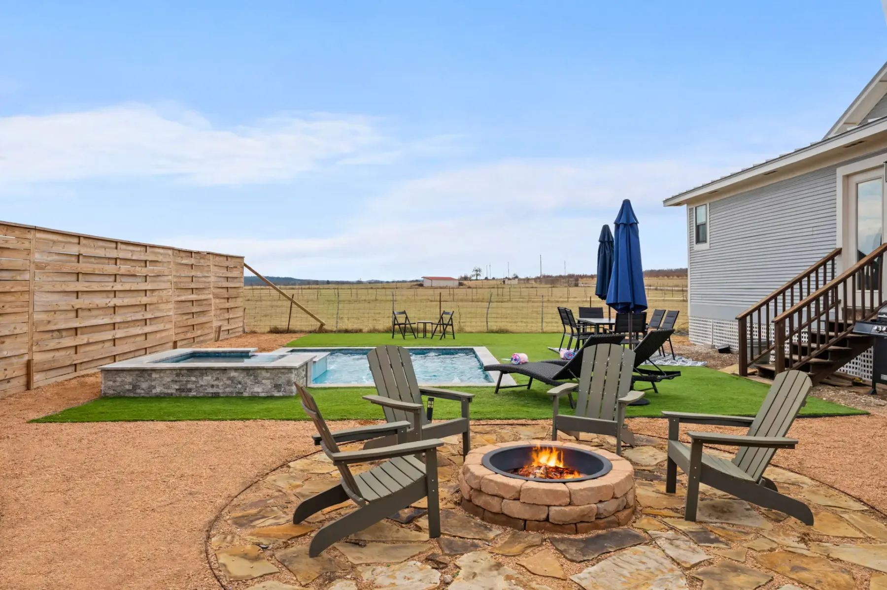 Backyard with a patio, fire pit surrounded by chairs, pool, hot tub, and outdoor furniture, set against a wooden fence and clear sky.