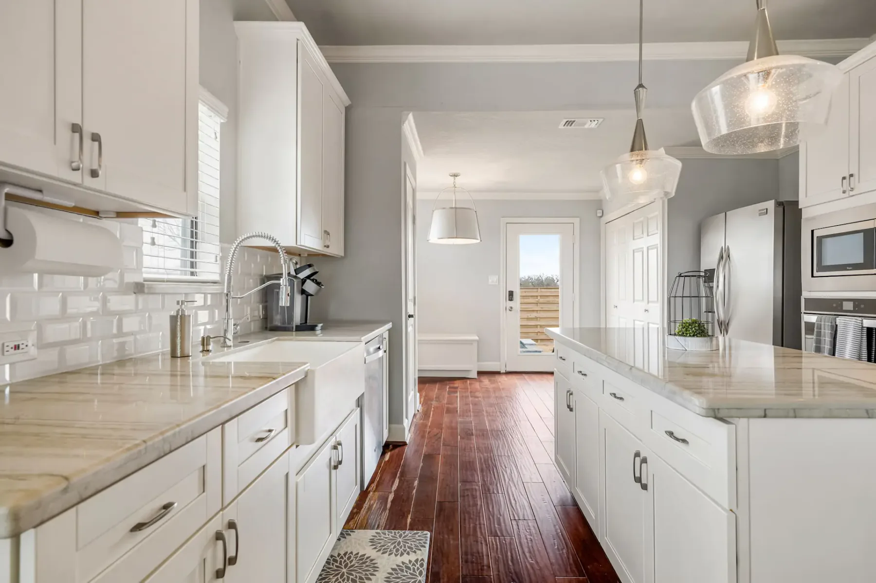 Modern kitchen with white cabinets, marble countertops, stainless steel appliances, and hardwood floors, leading to a door with natural light.