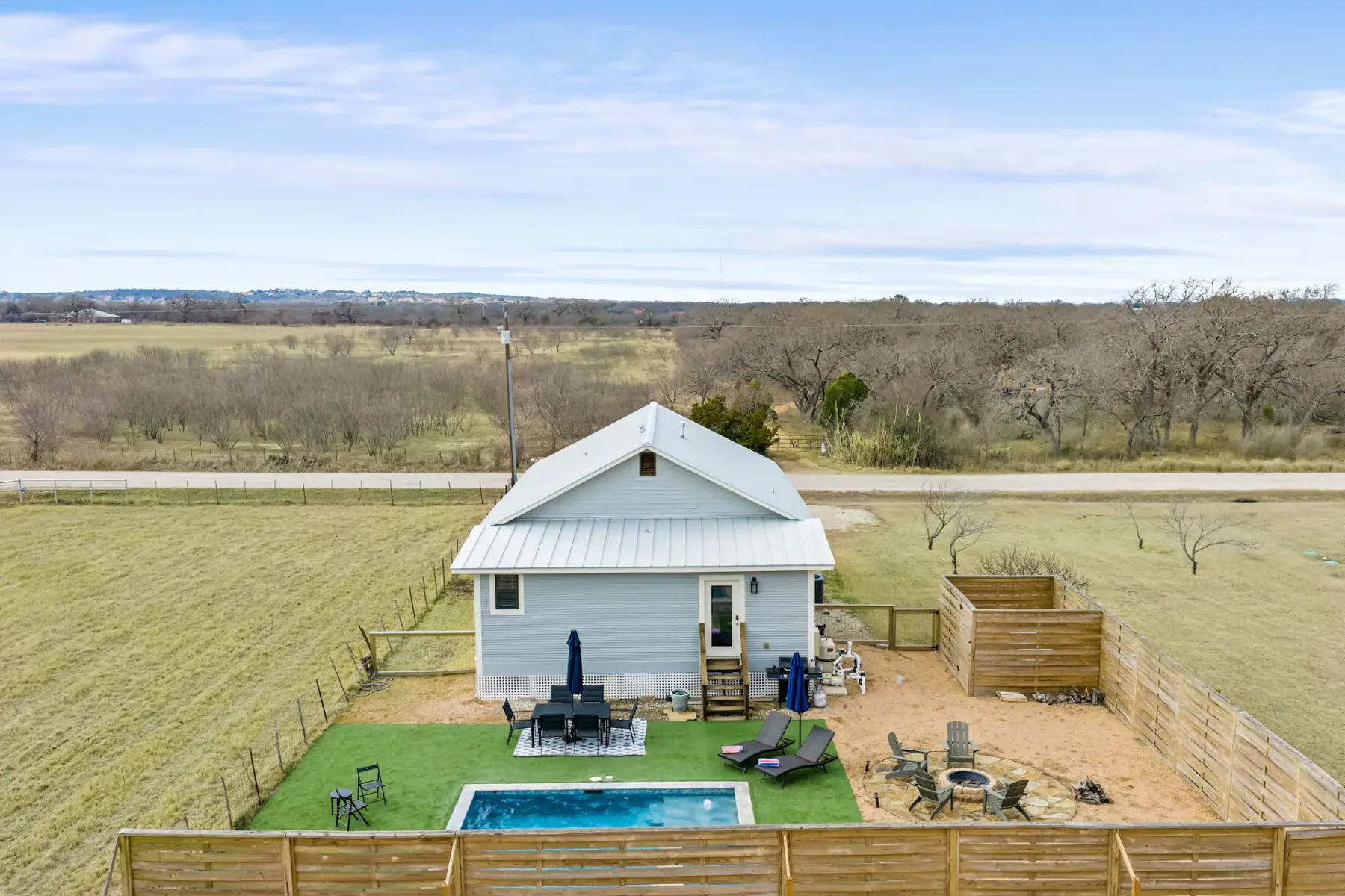 A white house with a fenced yard, a small pool, and open fields beyond.