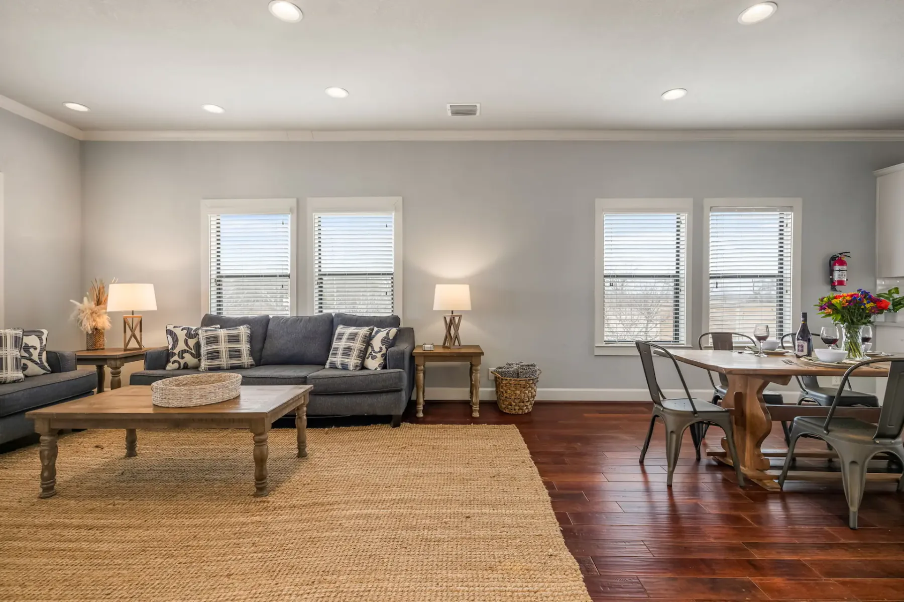 A cozy living and dining area with a sofa, coffee table, dining table with chairs, and large windows. Decor includes lamps and a woven rug.