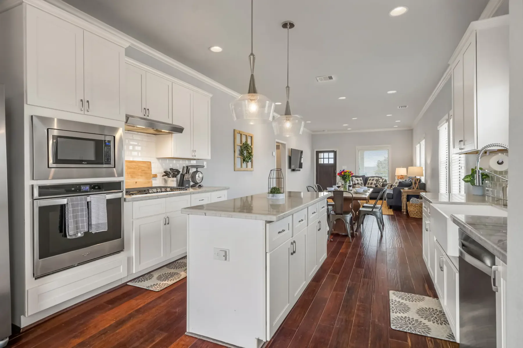 Modern kitchen with white cabinets, stainless steel appliances, marble island, pendant lights, and wooden flooring leading to a cozy living area.