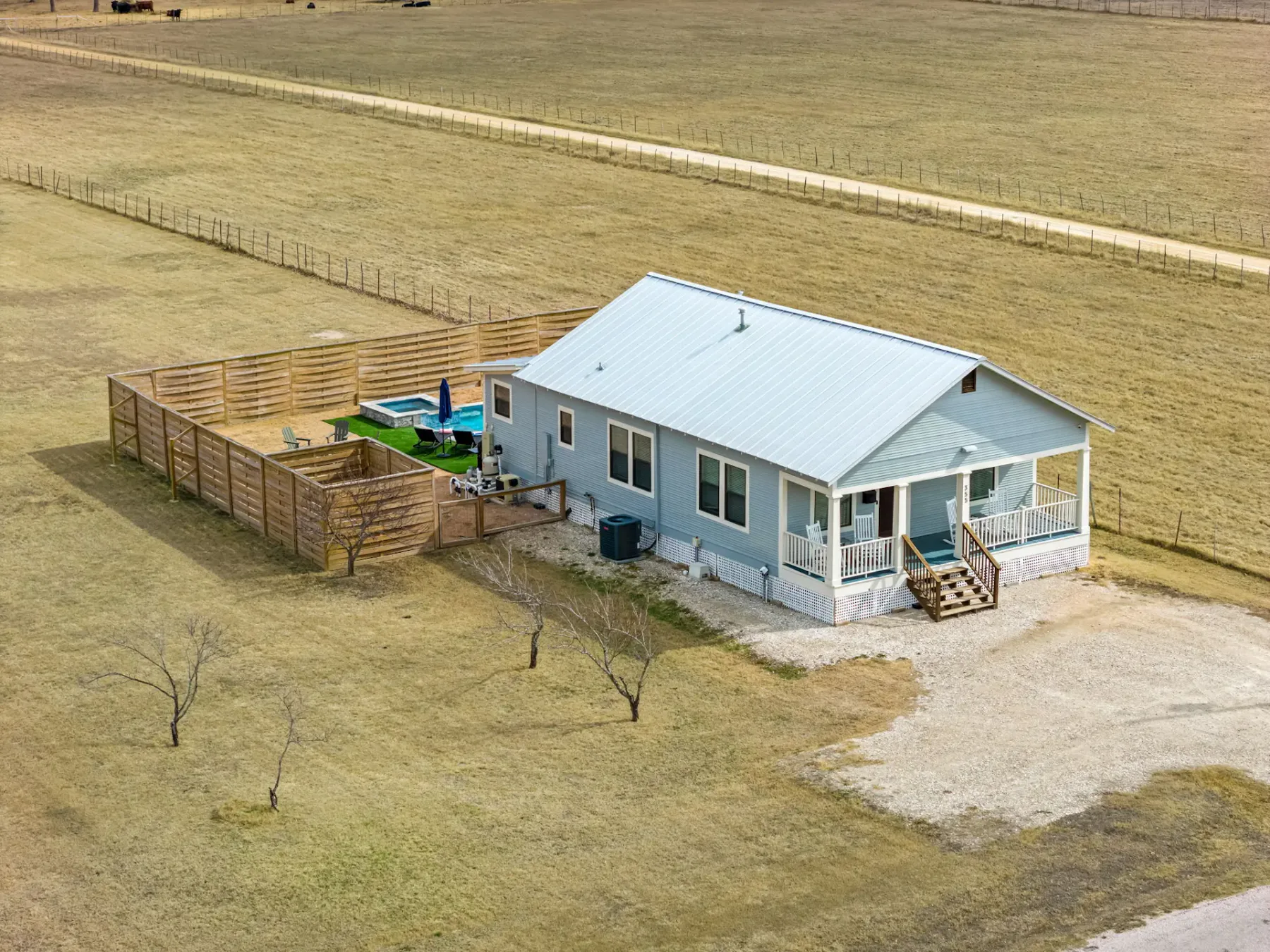 A small, light blue house with a porch, fenced backyard, and open field surroundings. It features a pool and seating area in the backyard.